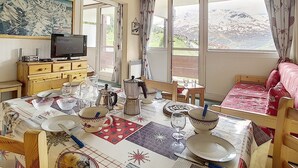 A cozy room with a dining table set for two, a television, and a view of a snowy landscape through large windows. - Spacious ski-in/ski-out apartment with balcony (Les Menuires)