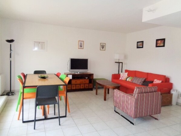 A living room with a red sofa, a wooden coffee table, a TV on a stand, a dining table with colorful chairs, and a floor lamp.