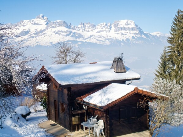 A wooden cabin with a snow-covered roof, a chimney, and a small patio with chairs.