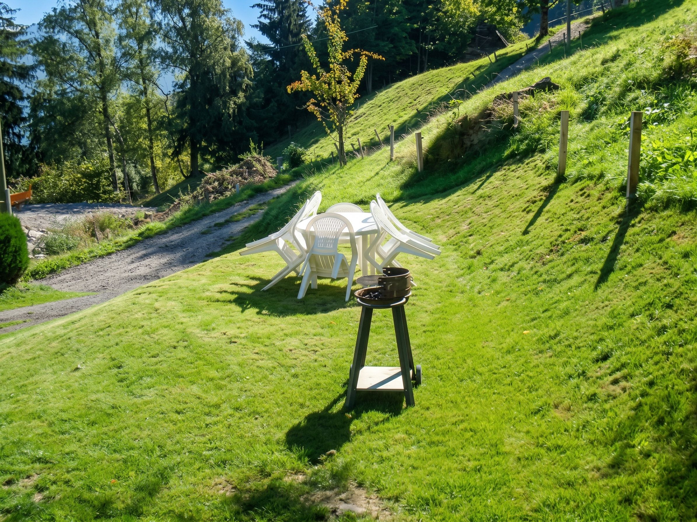 A grassy outdoor area with a white table and chairs, a metal grill, and a dirt path.