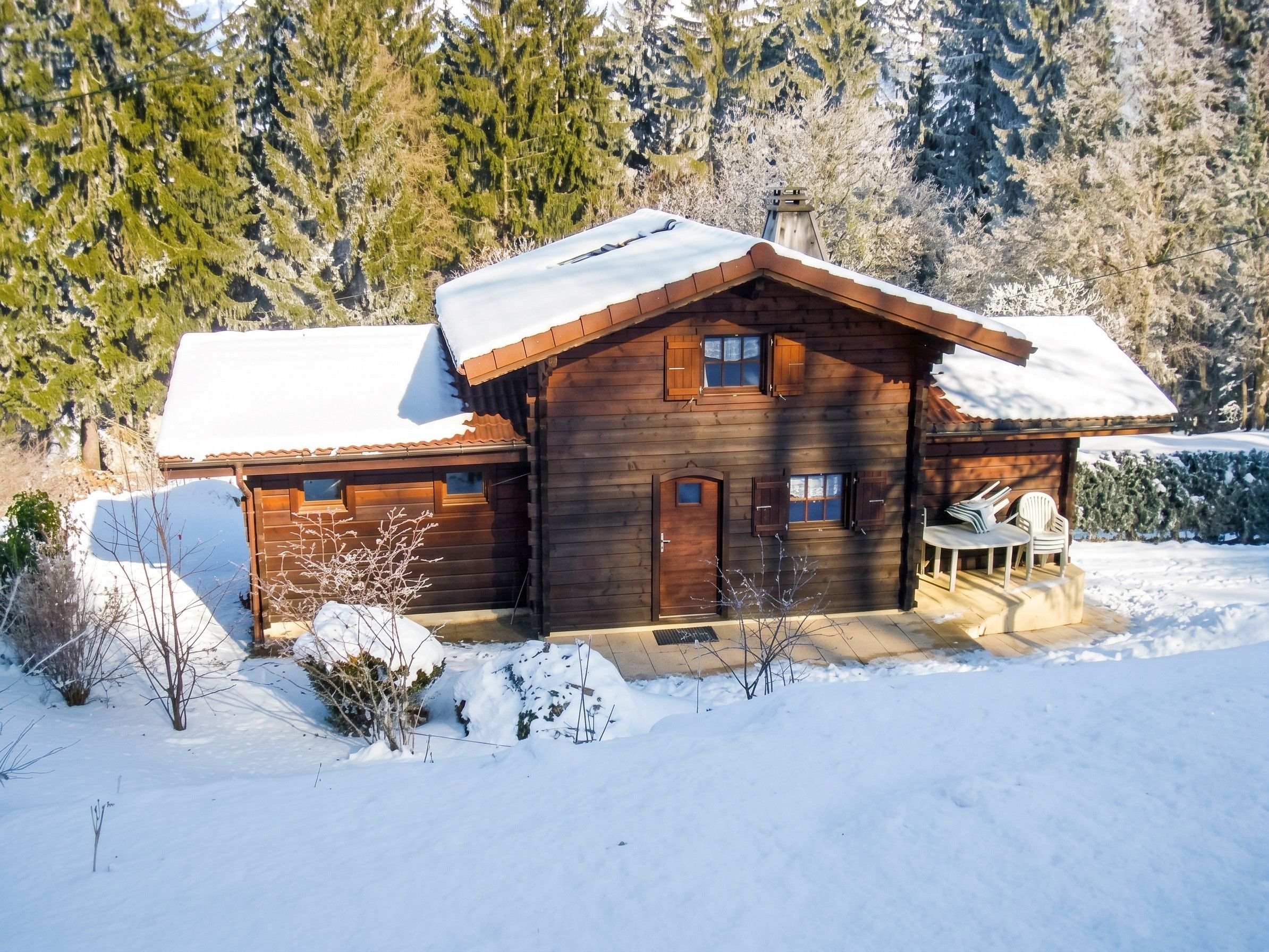 A wooden cabin with a snow-covered roof, a front porch with chairs, and a garage.