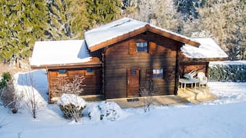 A wooden cabin with a snow-covered roof, a front porch with chairs, and a garage.