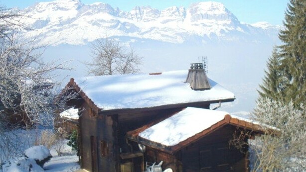 A rustic cabin with a snow-covered roof, a small patio with a table and chairs, and a mountain backdrop.
