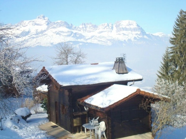 Una cabaña rústica con techo nevado, un pequeño patio con mesa y sillas, y montañas al fondo.