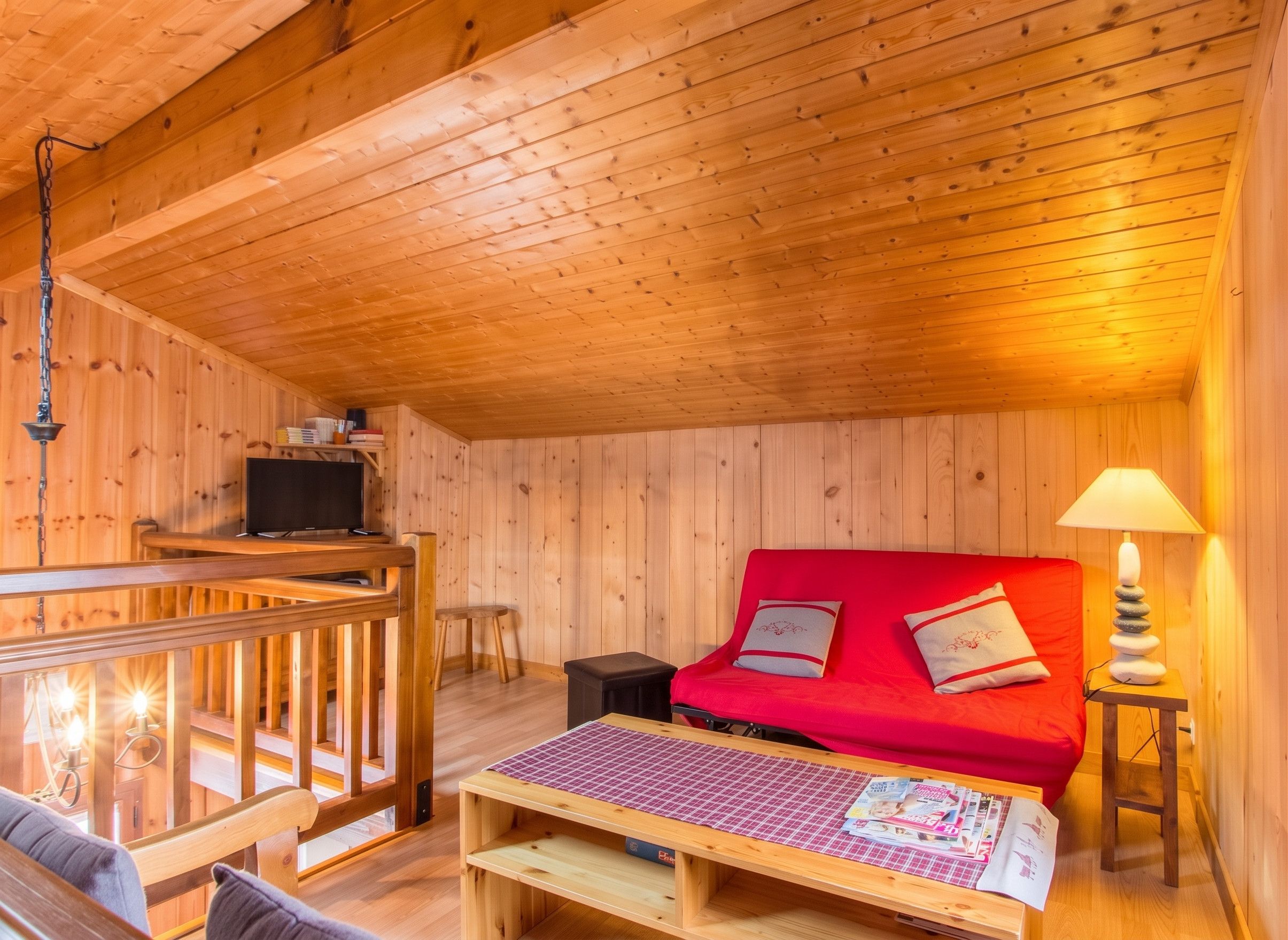 A cozy cabin interior with a wooden ceiling, a red sofa, a small table with magazines, and a lamp on a bedside table.