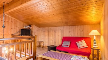A cozy cabin interior with a wooden ceiling, a red sofa, a small table with magazines, and a lamp on a bedside table.