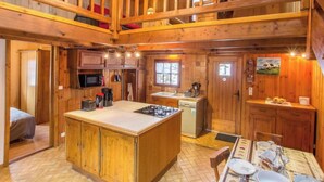 A wooden cabin with a central island kitchen, a dining area, and a loft bedroom.