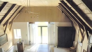 Ein Dachzimmer mit hoher Decke, Holzbalken, einem großen Tisch, Bänken und Blick durch eine Tür.