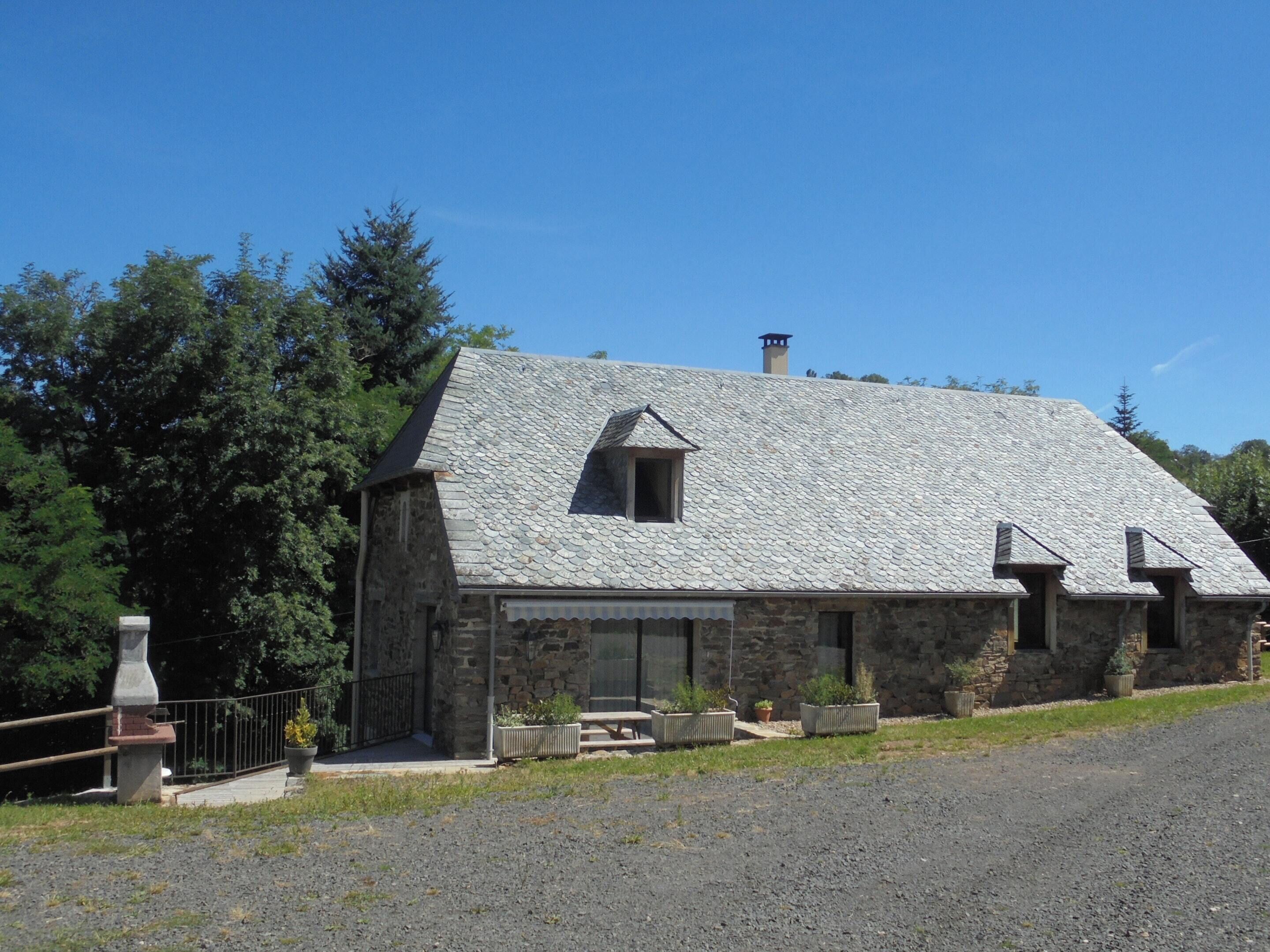 Una casa de piedra con techo inclinado y chimenea, rodeada de árboles y un camino de entrada de grava.