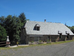 Ein Steinhaus mit einem geneigten Dach und Schornstein, umgeben von Bäumen und einer Schotterauffahrt.