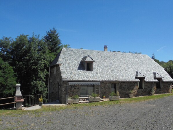 Una casa de piedra con techo inclinado y chimenea, rodeada de árboles y un camino de entrada de grava.