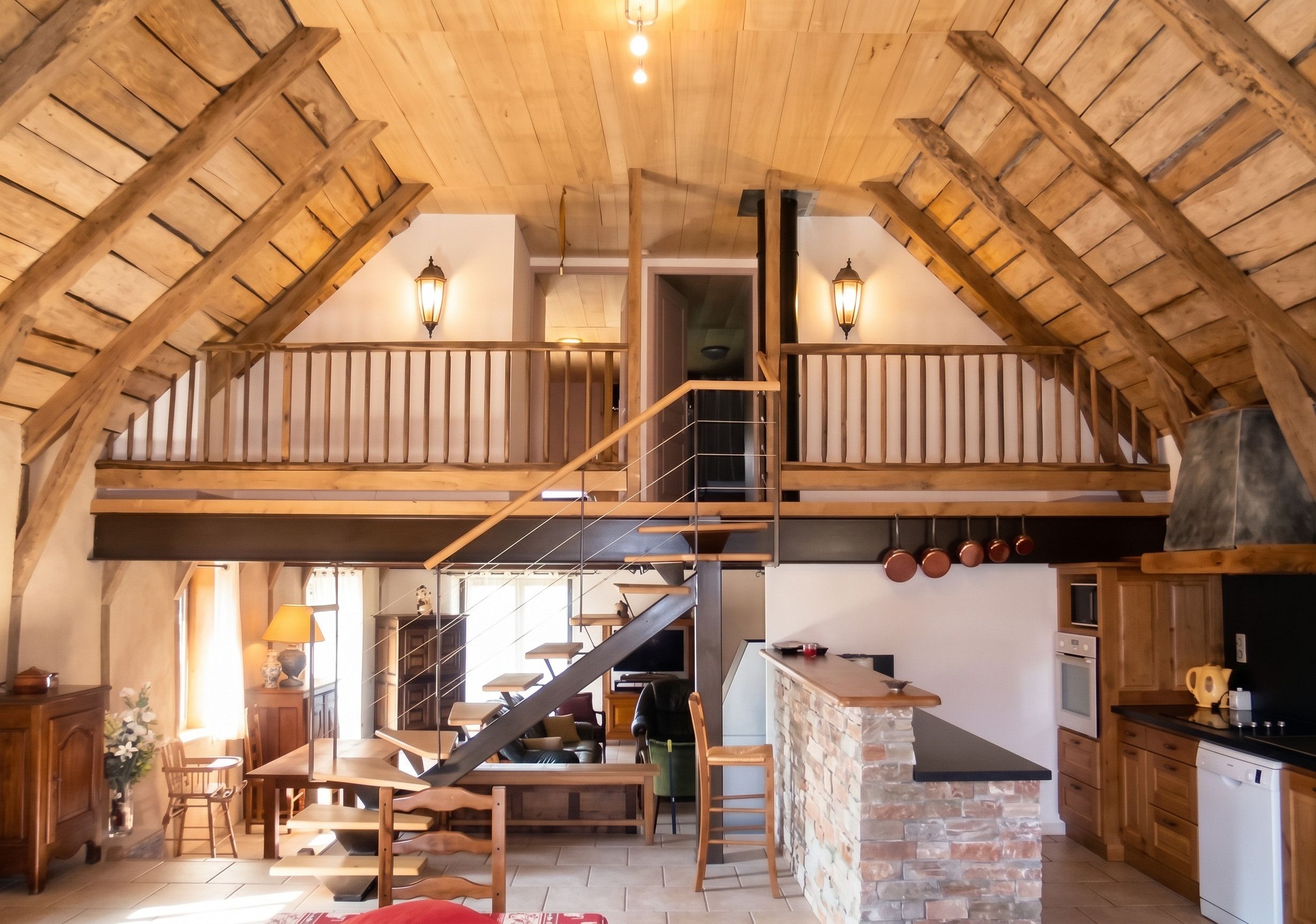 A spacious loft with a high ceiling, wooden beams, a mezzanine level, a kitchen area, and a staircase.