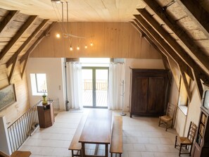 Ein geräumiges Dachzimmer mit Holzboden, einem Esstisch und einer Bank. Es gibt ein großes Fenster mit Vorhängen, eine Heizung und einen Kleiderständer.