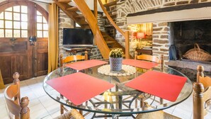 A cozy living room with a stone wall, wooden beams, a fireplace, a TV, and a round glass table with red placemats.
