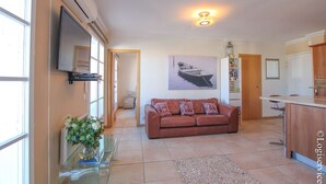 A spacious living area with a brown leather sofa, a flat-screen TV mounted on the wall, a painting of a boat, and a kitchen area with a wooden counter and chairs. - Lovely renovated flat for 4 persons with AC and mooring (PORT GRIMAUD)