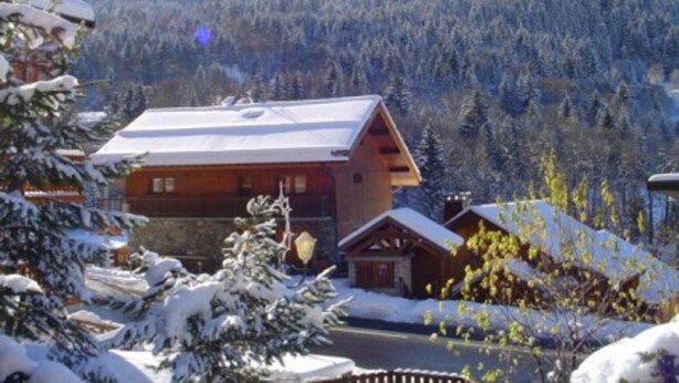 Uma cabana de madeira com telhado inclinado, cercada por árvores cobertas de neve e montanhas ao fundo.