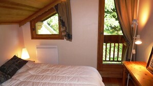 Quarto com cama, mesa de cabeceira, abajur, janela com cortinas e vista para o verde lá fora.