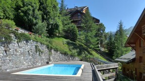 Um deck de madeira com uma piscina retangular, ladeado por muros de pedra e vegetação densa.