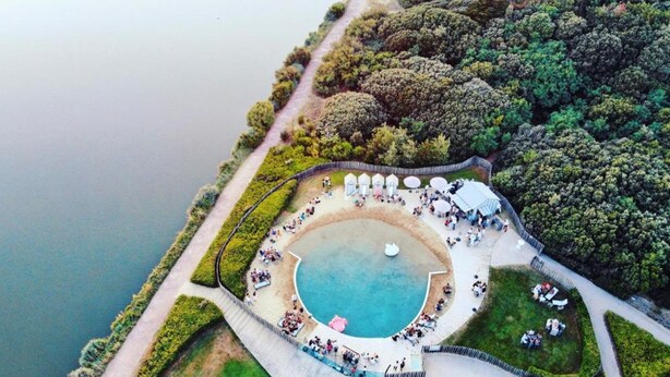 An aerial view of a circular pool surrounded by a deck with lounge chairs and umbrellas, set in a landscaped area with trees and a pathway leading to the pool. - Elegant Apartment with Garage – 400m from Beach (Les Sables-d'Olonne)