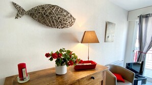 A living room with a wooden cabinet, a red candle, a potted plant with red flowers, a lamp, and a fish wall decoration. - Elegant Apartment with Garage – 400m from Beach (Les Sables-d'Olonne)
