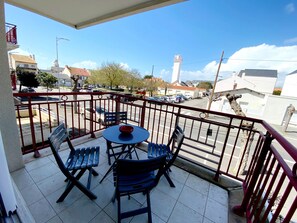Un balcon avec une table et des chaises, une vue sur une rue où circulent des voitures, et un immeuble doté d’une tour.