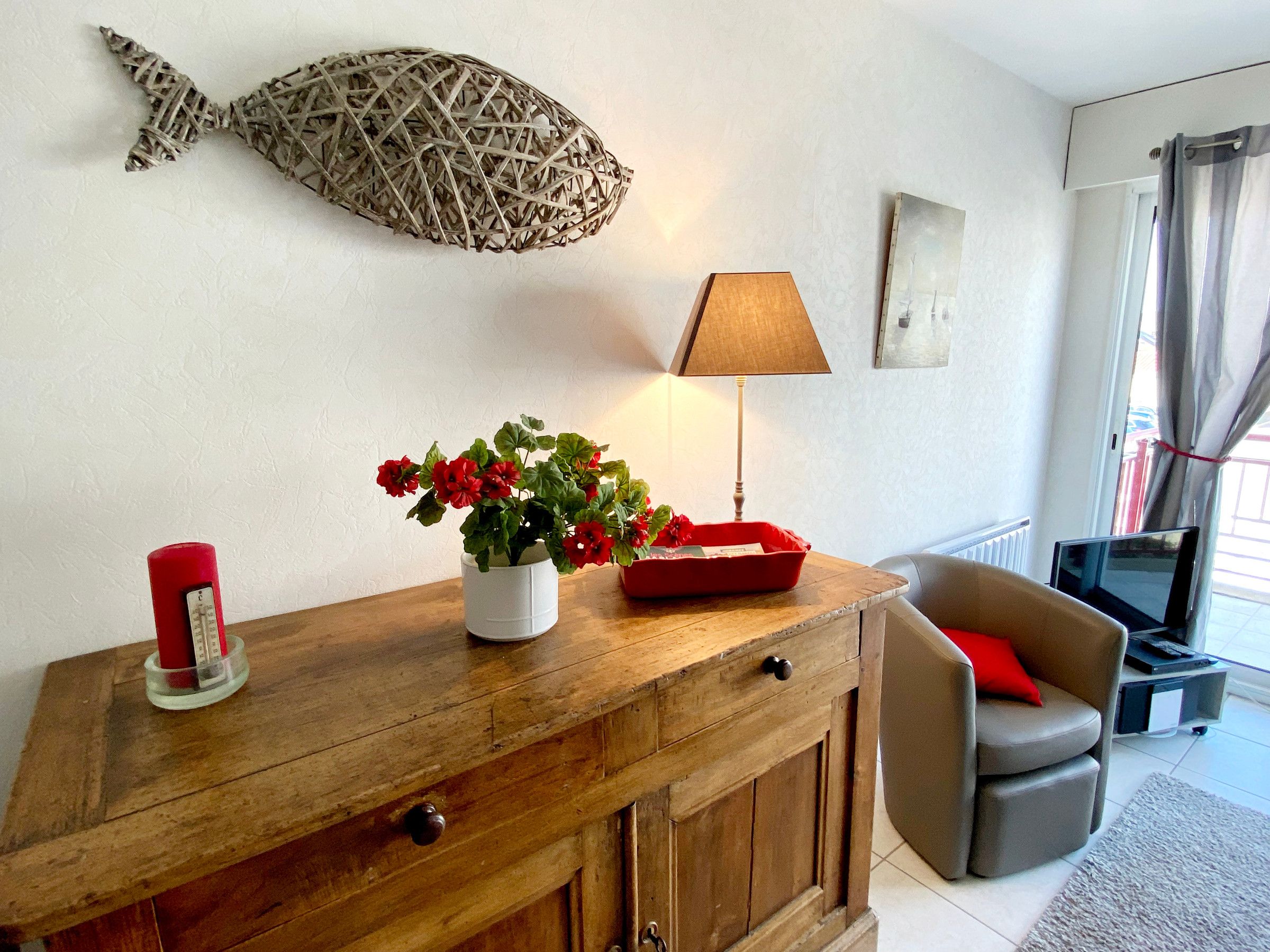 Ein Wohnzimmer mit einem Holzkabinett, einer roten Blumenvase, einer Lampe, einer Fisch-Wanddekoration, einem grauen Sofa und einem roten Kissen.