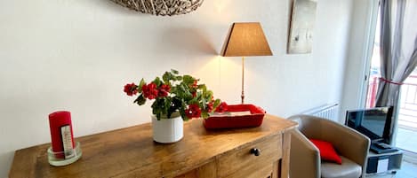 A living room with a wooden cabinet, a red flower vase, a lamp, a fish wall decoration, a grey sofa, and a red cushion.