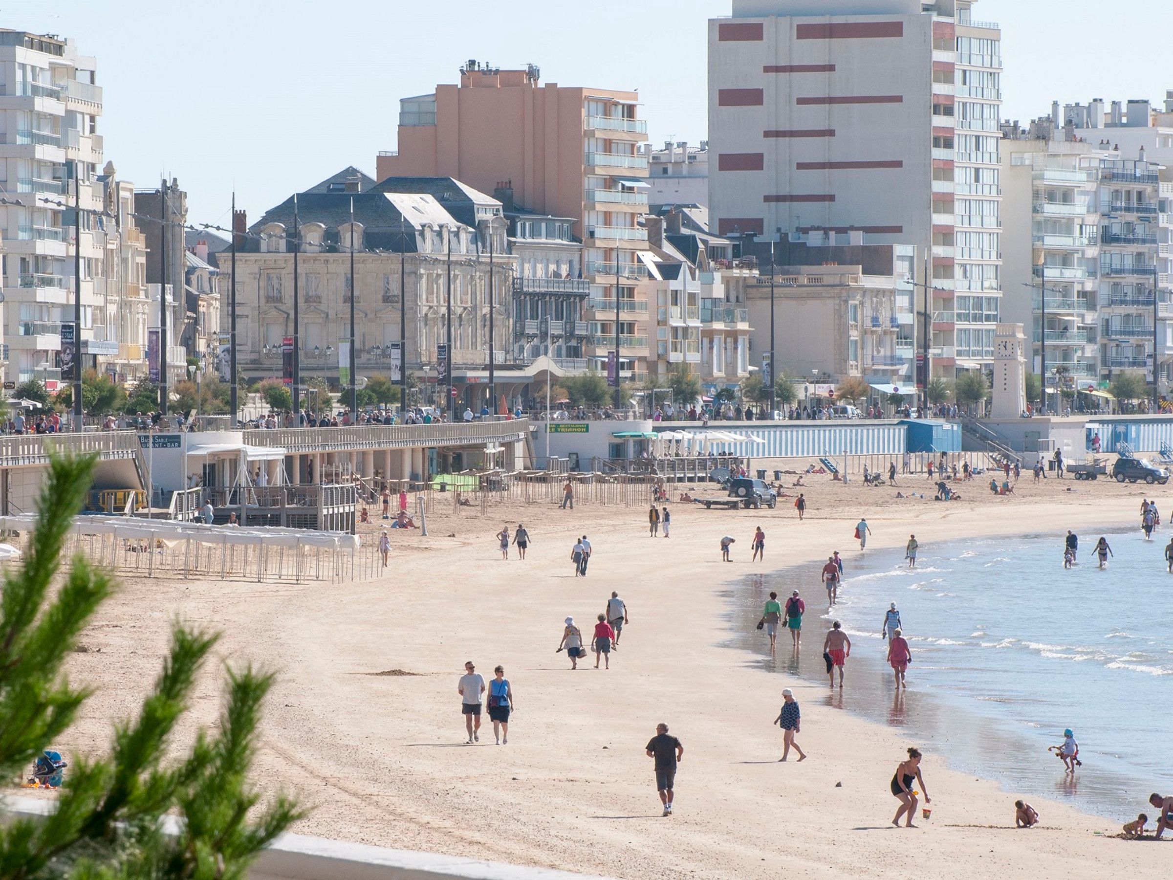 Una spiaggia con persone che passeggiano e giocano, edifici sullo sfondo e un cielo sereno.