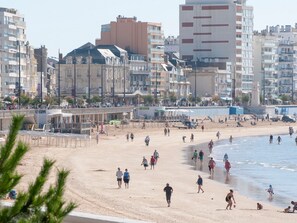 Une plage où l’on voit des gens se promener et jouer, des bâtiments en arrière-plan et un ciel dégagé.