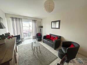 Un salon avec un canapé, un fauteuil et une petite table. Il y a un balcon avec vue sur l’extérieur.