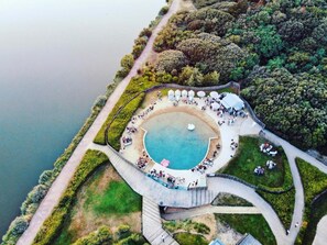 Une vue aérienne d’une piscine circulaire entourée d’une terrasse agrémentée de chaises longues et de parasols, située dans un espace paysager avec des arbres et un sentier menant à la piscine.
