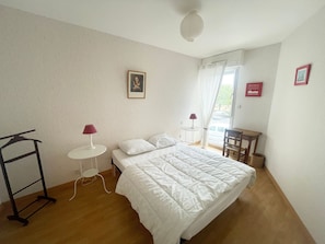 Un dormitorio con cama, una mesita, una silla, perchero y ventana con cortinas.