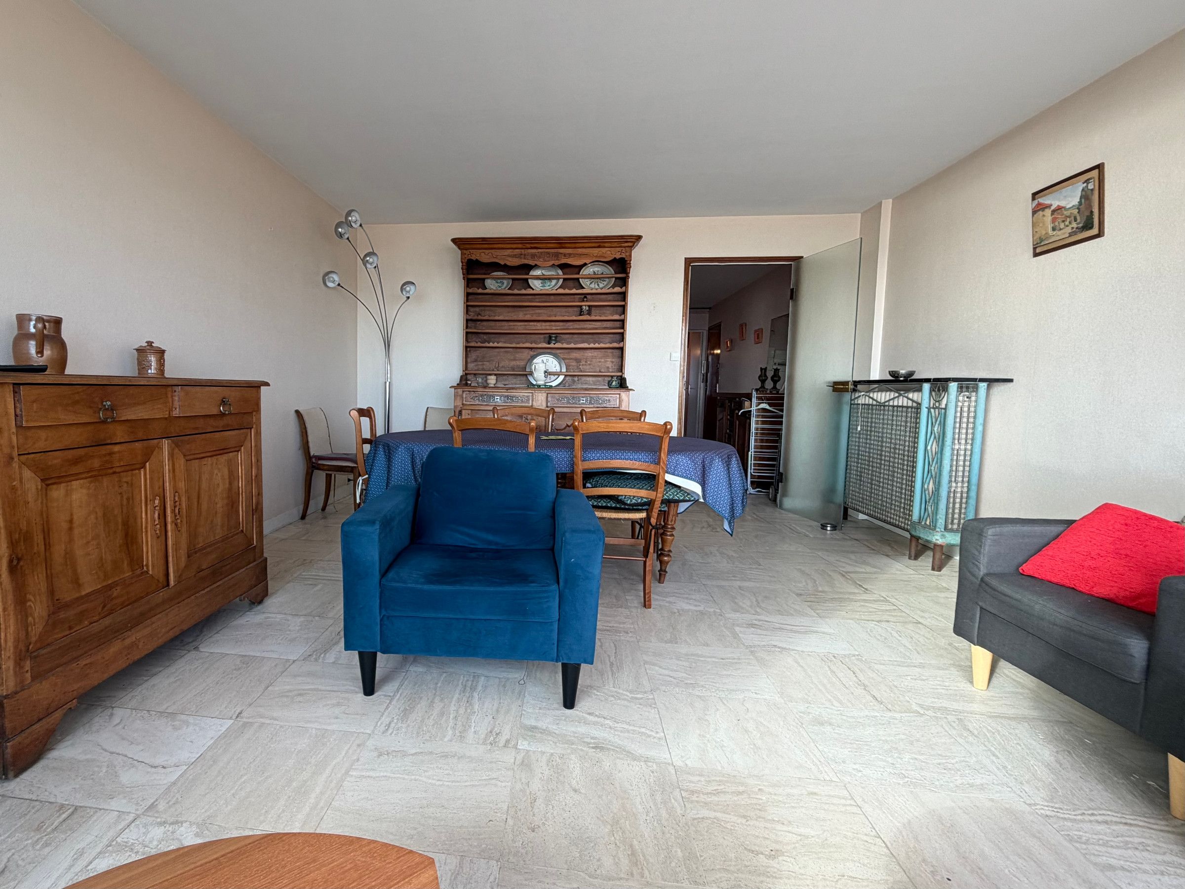 Una sala de estar con un sillón azul, un aparador de madera, una mesa de comedor con sillas y un sofá gris con un cojín rojo.