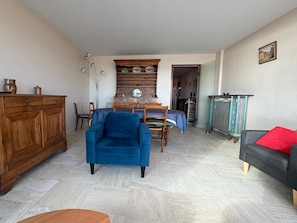 A living room with a blue armchair, wooden cabinet, dining table with chairs, and a grey sofa with a red pillow.