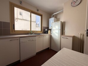 A compact kitchen with a dishwasher, a refrigerator, a coffee maker, and a window with a view of buildings outside.