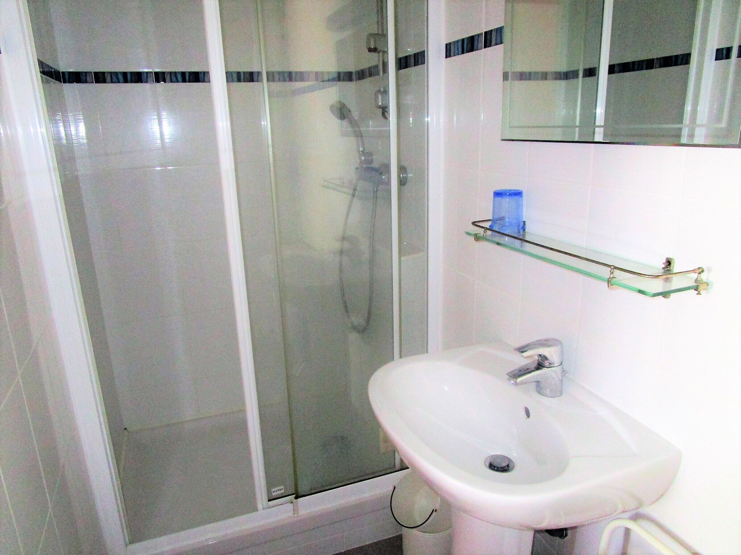 Un baño con ducha envuelta en cristal, un lavamanos blanco y un estante de vidrio sobre el lavamanos.