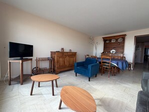 A living room with a television, wooden furniture, a blue armchair, and a dining area with a table and chairs.