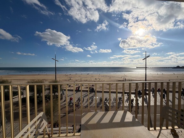 A beach with a clear sky, a few clouds, and a sun shining. There are people walking and cycling on the beach and a fence in the foreground.