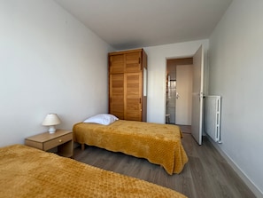 A bedroom with two beds, a wooden wardrobe, a radiator, and a nightstand with a lamp.