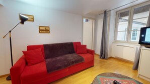 Un canapé rouge avec un plaid marron, un lampadaire, un téléviseur et une fenêtre avec des rideaux.