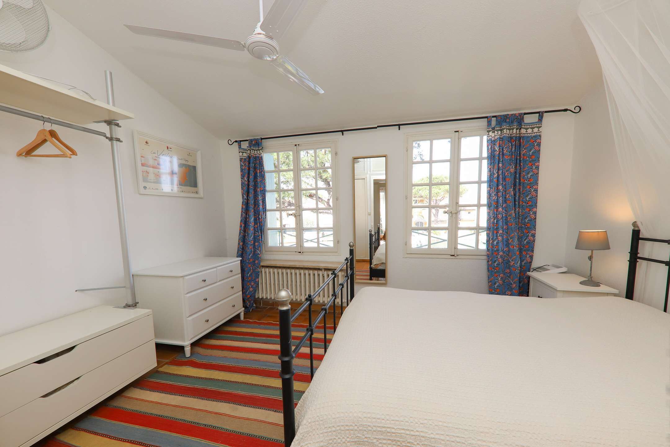 A bedroom with a bed, a dresser, a radiator, a ceiling fan, and a window with curtains.