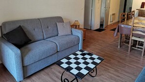A living room with a grey sofa, a checkered coffee table, and a dining area with a wooden table and chairs. - Comfortable apt for 8, 2 bdr, terrace, laundry, near slopes (La Mongie)