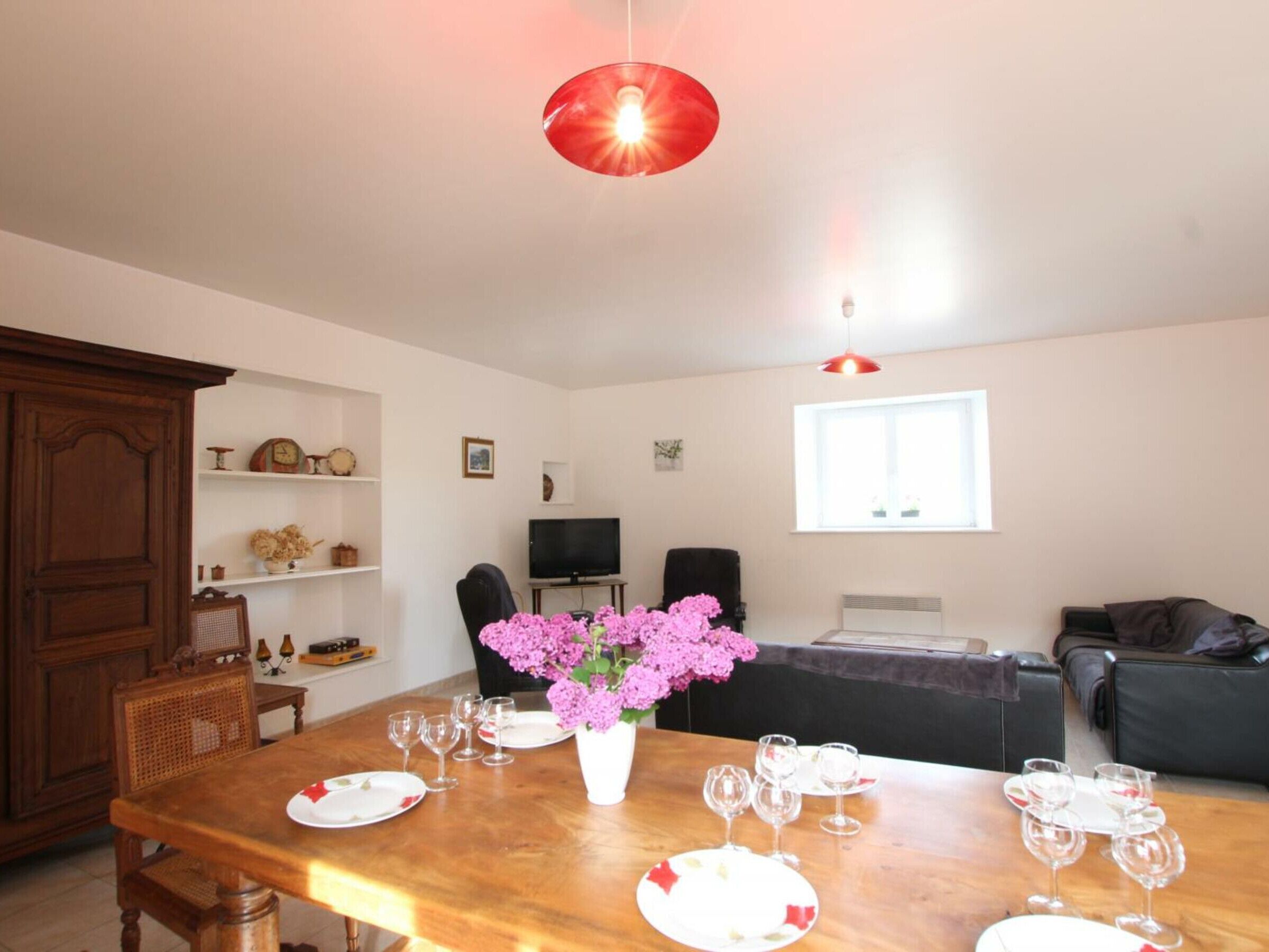 A dining area with a wooden table set for four, a vase with pink flowers, and a television in the background.