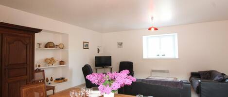 A dining area with a wooden table set for four, a vase with pink flowers, and a television in the background.