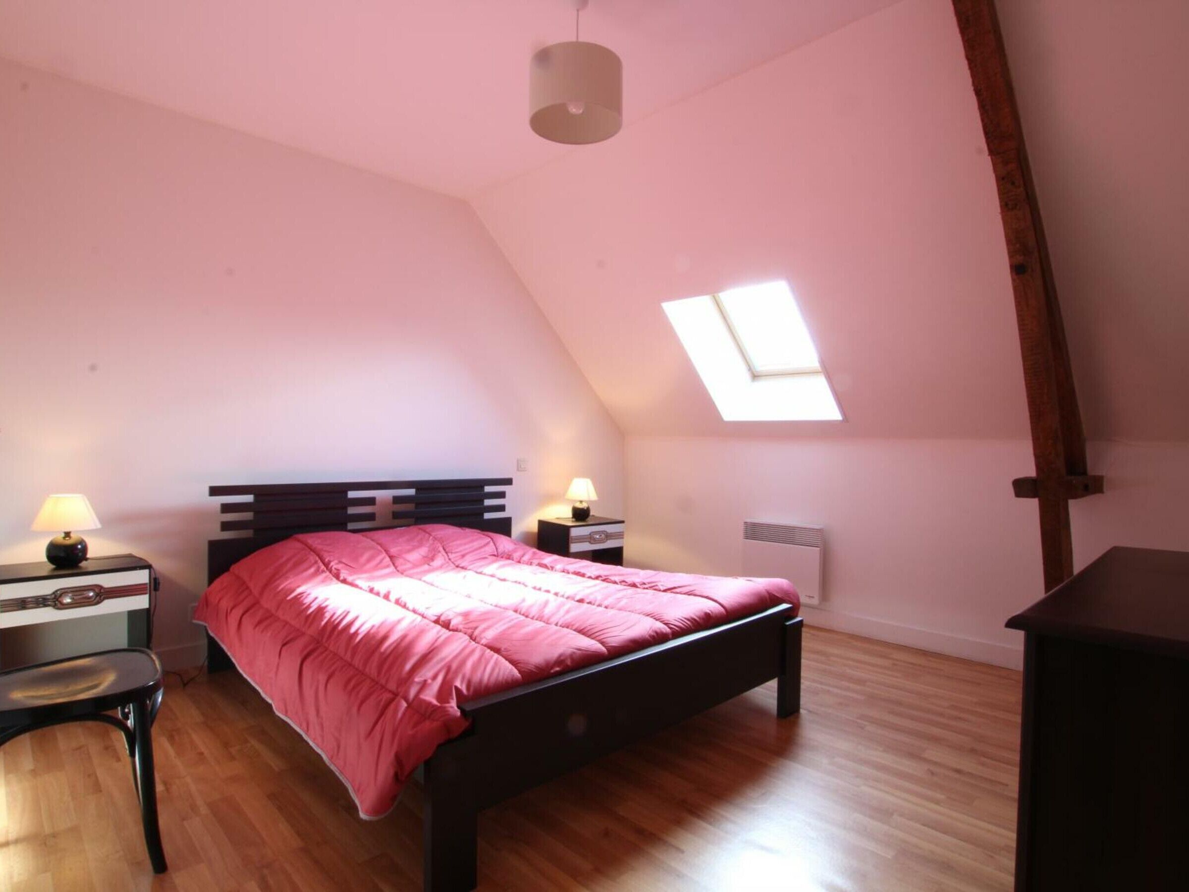 A bedroom with a sloped ceiling, a skylight, a bed with a pink blanket, two bedside tables with lamps, and a wooden chair.