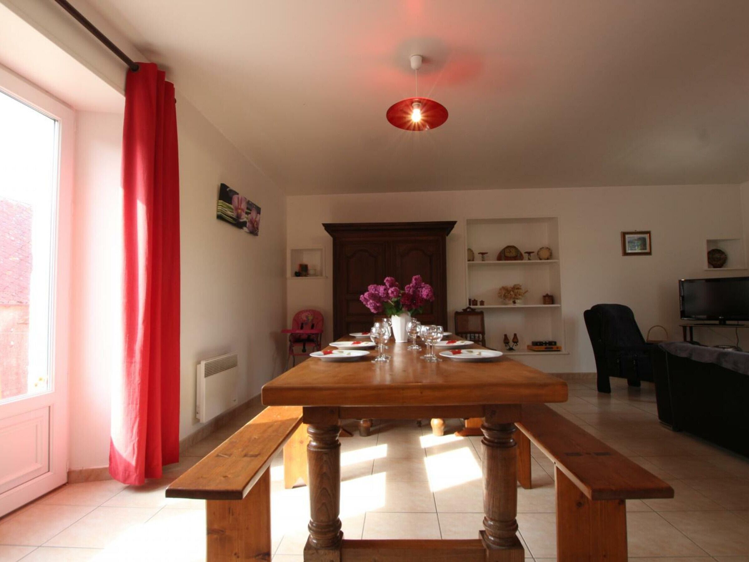 Un coin repas avec une table en bois dressée pour quatre personnes, un vase de fleurs et un téléviseur en arrière-plan.