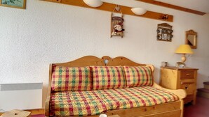 A cozy room with a wooden bed, a small table with cups, a wooden dresser, and a wall clock. - Bright ski-in/ski-out duplex, balcony, 4 ppl (Les Menuires)