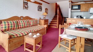 A cozy living room with a wooden sofa, a small wooden table with cups, a staircase leading to a kitchen area with wooden cabinets and a microwave. - Bright ski-in/ski-out duplex, balcony, 4 ppl (Les Menuires)