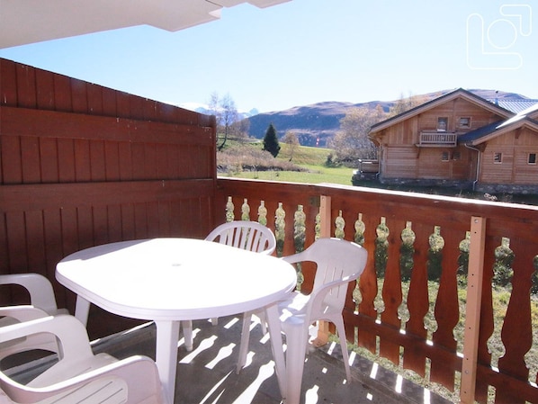 Um deck de madeira com uma mesa e cadeiras brancas, com vista para uma paisagem rural com casas e montanhas.