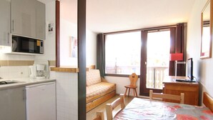 A compact kitchenette with a microwave, coffee maker, and a small refrigerator. There is a bed with a patterned bedspread, a wooden chair, and a desk with a computer. A large window provides natural light and a view of the outside. - Cozy 2-Room Apartment for 6, Near Slopes & Shops, Huez (Huez)